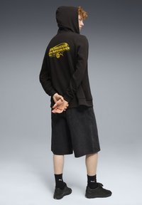 Sort hættejakke med gult "Borussia Dortmund" logo på ryggen, parret med sorte denimshorts. Sorte sokker og sneakers fuldender looket.