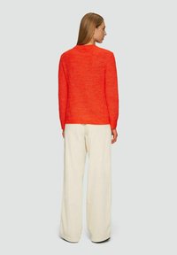 Oranges Strickpullover mit langen Ärmeln, rundem Halsausschnitt und geripptem Textil, kombiniert mit weißen, weit geschnittenen Hosen, die von hinten einen lässigen Sitz zeigen.