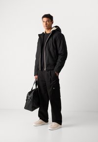 Giacca nera isolante con cappuccio, foderata in pelliccia beige, chiusura con zip, abbinata a pantaloni cargo neri e sneakers beige chiaro. Borsa nera in mano.