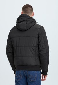 Black padded jacket with a hood, featuring quilted texture and ribbed cuffs. The jacket has a smooth, matte finish and a relaxed fit.