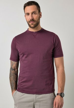 Mężczyzna z brodą i wytatuowanym ramieniem, ubrany w bordową koszulkę z krótkim rękawem i beżowe spodnie, stojący na tle jednolitego szarego tła.