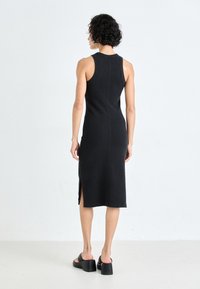 Εφαρμοστό μαύρο αμάνικο φόρεμα με ψηλό λαιμό, λεπτομέρεια ραφής στην πλάτη, πλαϊνό άνοιγμα και ριγωτή υφή, συνδυασμένο με χοντρά μαύρα σανδάλια.