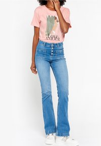 T-shirt rosa chiaro con un design grafico e testo nero, abbinata a jeans blu a vita alta con cinque bottoni e due tasche frontali.