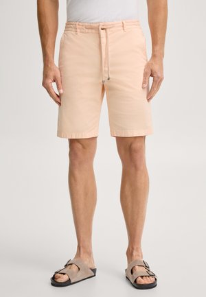 Mann trägt hellpfirsichfarbene knielange Shorts mit Kordelzug und beige Sandalen, steht vor einfachem Hintergrund.