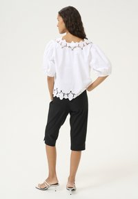 Camicia di cotone bianca con maniche a sbuffo, rifinita in pizzo sul collo e sull'orlo, abbinata a pantaloni neri al ginocchio. Calzature semplici visibili.