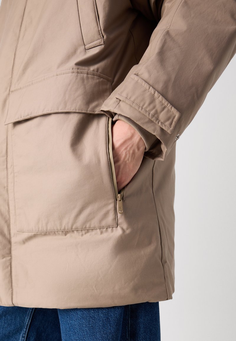 Beige geïsoleerde jas met een ritszijdezak en een grote klepzak, met een gladde textuur en een iets losse pasvorm.