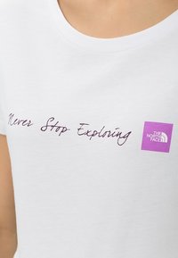 Bílý bavlněný tričko s kurzivním textem "Nikdy nepřestávej objevovat" v purpurové barvě a čtvercovým purpurovým logem The North Face vpravo.