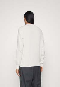 Pull crème clair en tricot doux, col rond, et poignets et ourlet côtelés. Porté avec un pantalon ample gris foncé.