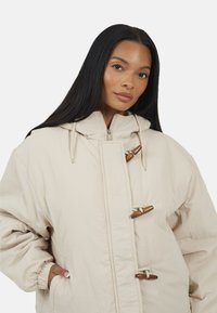 Beżowa kurtka puchowa z kapturem, z drewnianymi zapięciami w formie suwaków, elastycznymi mankietami i zamkiem błyskawicznym z przodu. Gładka, pikowana faktura.