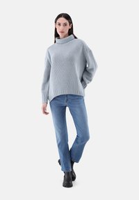 Hellgrauer gestrickter Pullover mit hohem Kragen, kombiniert mit blauen geraden Jeans und schwarzen klobigen Stiefeln, vor einem neutralen Hintergrund.