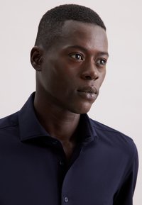 Camicia blu navy con bottoni, caratterizzata da una texture morbida, un colletto classico e un design aderente. Il tessuto appare liscio e brillante.