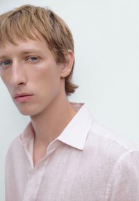 Lichtroze linnen shirt met een puntige kraag, gestructureerde stof en zachte plooien. Model toont een neutrale uitdrukking tegen een effen achtergrond.