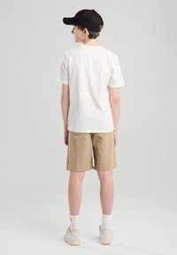 Valkoinen puuvillapaita, beige shortsit ja valkoiset tennarit. Musta lippis ja valkoiset sukat näkyvissä. Keskity yksinkertaiseen, rentoon muotoiluun ja väri-eroihin.