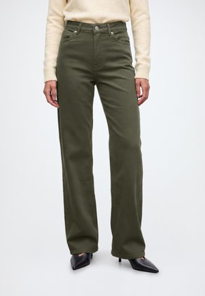 Femme portant un pantalon large taille haute vert olive, un pull crème et des talons pointus noirs, debout sur un fond blanc.