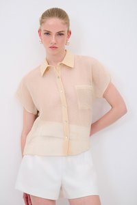 Camicia corta a maniche corte beige chiaro con un motivo a righe testurizzato, chiusura con bottoni e taschino sul petto, abbinata a pantaloni corti bianchi.