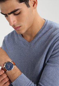 Pull bleu en V, tenant une montre avec un cadran bleu et un boîtier en acier inoxydable, bracelet en cuir marron et détails de chronographe.