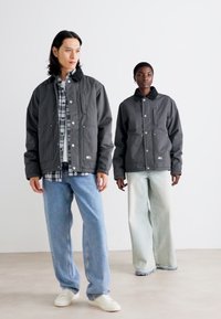 To modeller bærer mørkegrå jakker med trykknapper og lappelommer over ternede skjorter. De parrer dem med lyse jeans og hvide sneakers.