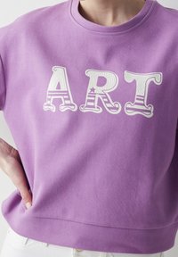 Lila sweatshirt i bomull med djärva, vita broderade bokstäver som stavas "ART" med randiga detaljer på bokstäverna. Justerbar passform.
