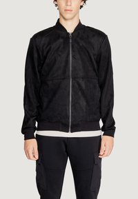 Giacca bomber nera, morbido tessuto scamosciato, chiusura con zip frontale, polsini e orlo a costine, cuciture dettagliate, indossata su una camicia chiara.