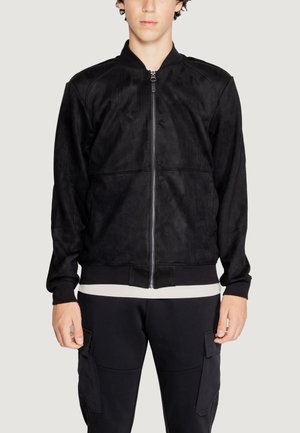 Chaqueta bomber negra, suave textura de ante, cierre delantero de cremallera, puños y dobladillo acanalados, costuras detalladas, llevada sobre una camisa clara.