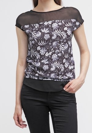 Femme portant un chemisier noir à motifs floraux avec un col en voile noir transparent et un pantalon noir, debout avec une main dans la poche.