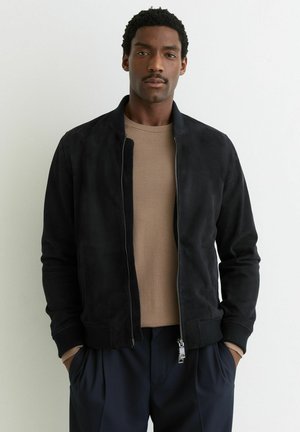 Man met kort krullend haar, gekleed in een zwarte jas met ritssluiting over een beige trui en donkere broek, staand tegen een effen witte achtergrond.