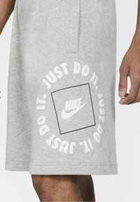 Shorts de sudadera Nike en gris con un gráfico circular blanco que presenta "JUST DO IT." y el logo de Nike en el centro. Textura suave, ajuste relajado.