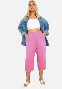 Vaaleanpunaiset lyhyet housut joustavalla vyötärönauhalla, yhdessä valkoisen lyhyen paidan ja sinisen farkkutakin kanssa. Henkilöllä on jalassaan valkoiset liukusandaalit.