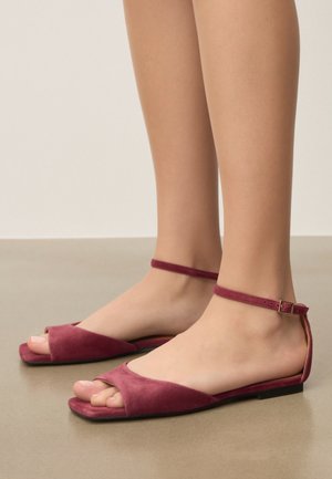 Noge v odprtih nizkih sandalih iz bordo rdečega semiša, z ozkimi trakovi okoli gležnjev in majhnimi zlatimi zaponkami, ki stojijo na gladki podlagi.
