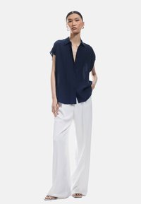 Camicia blu navy a maniche corte con bottoni e vestibilità rilassata, abbinata a pantaloni bianchi a gamba larga. Tessuto leggero con una texture liscia.