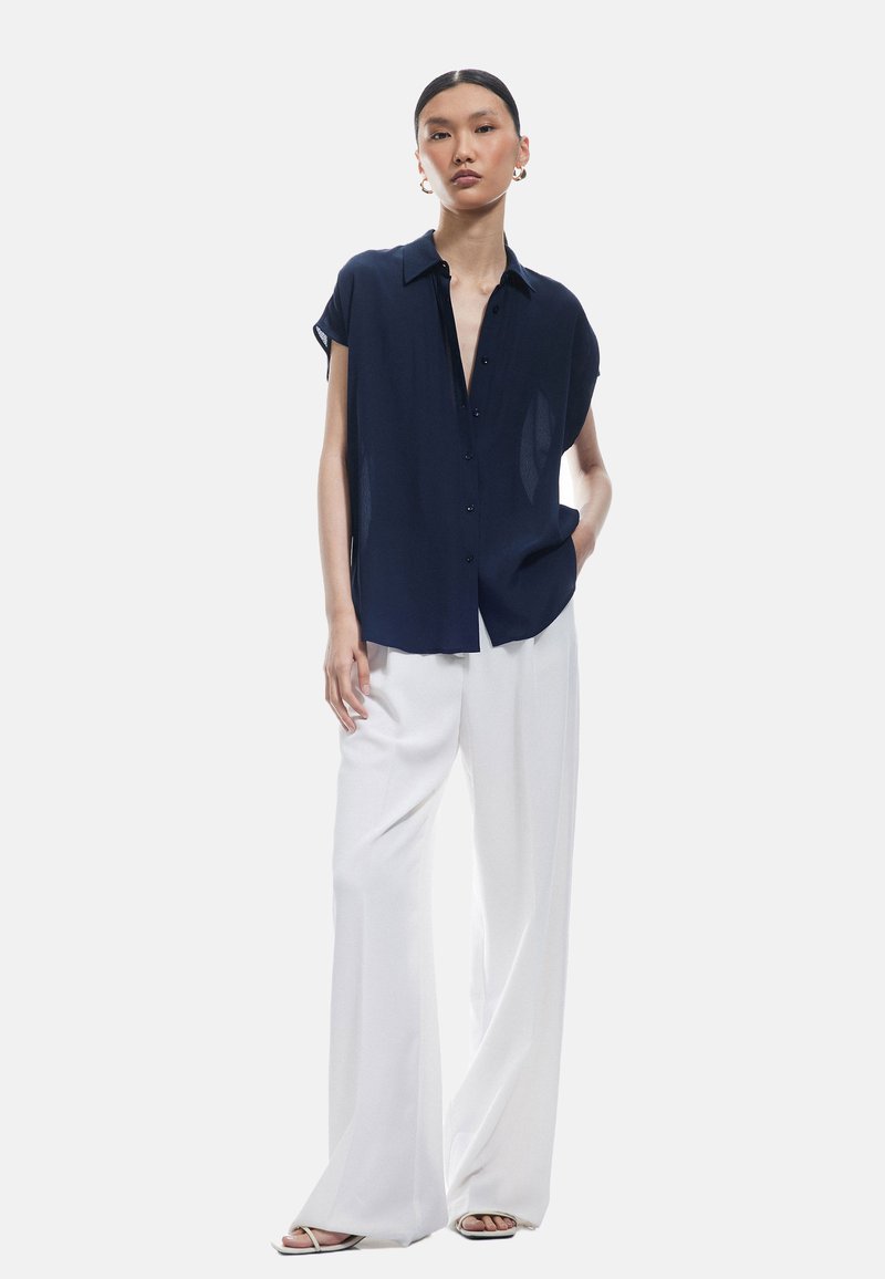 Camicia blu navy a maniche corte con bottoni e vestibilità rilassata, abbinata a pantaloni bianchi a gamba larga. Tessuto leggero con una texture liscia.