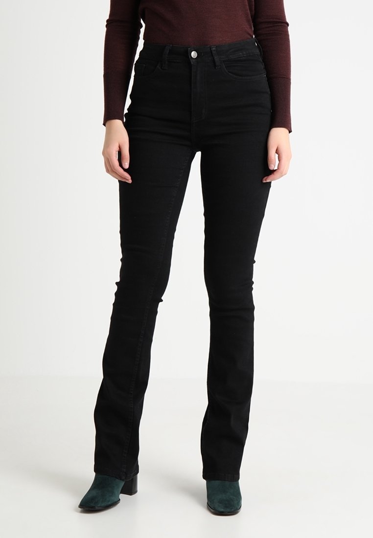 Femme portant un jean noir slim taille haute et des bottines en daim bleu sarcelle foncé, debout sur un fond blanc.