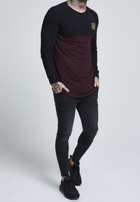 Man med tatueringar på halsen och händerna, iklädd en svart och vinröd långärmad tröja, svarta smala jeans och svarta sneakers, stående med händerna i fickorna.