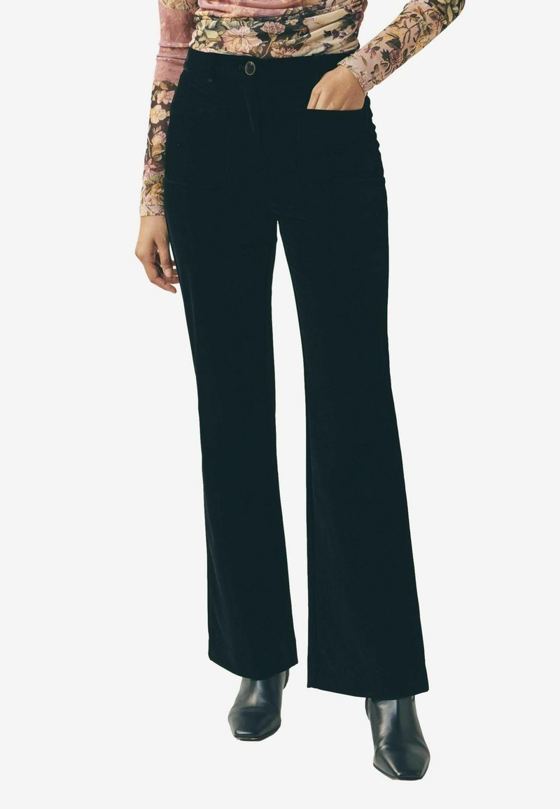 Kobieta ubrana w czarne szerokie spodnie, kwiecistą bluzkę z długim rękawem i czarne botki na obcasie, z jedną ręką w kieszeni, stojąca prosto.