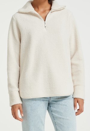 Person, die einen cremefarbenen Sherpa-Fleece-Pullover mit Halbreißverschluss und hellblaue Jeans vor schlichtem Hintergrund trägt.