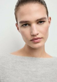 Suéter gris con textura de canalé y escote amplio. La modelo tiene el cabello corto y peinado hacia atrás, y un maquillaje natural, con un fondo neutro.