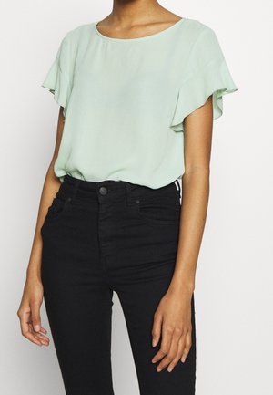 Woman wearing a light green short-sleeve blouse with ruffled sleeves tucked into high-waisted black pants.