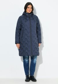 Cappotto imbottito blu navy con cappuccio, caratterizzato da un motivo a zigzag, chiusura frontale con zip e due tasche laterali. Indossato sopra un denim casual.