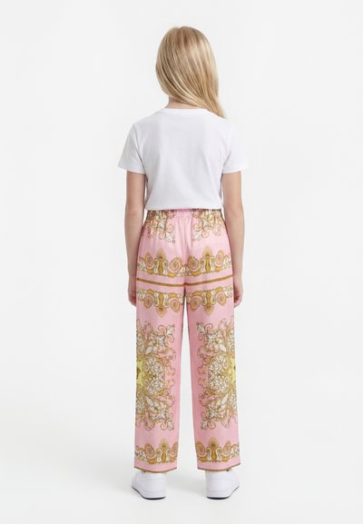 Enfant aux longs cheveux blonds de dos, portant un t-shirt blanc, un pantalon large rose avec des motifs dorés ornés, et des baskets blanches.