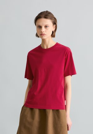 Jong persoon met kort haar, een effen rood t-shirt en een bruine rok, staand tegen een lichtgrijze achtergrond.