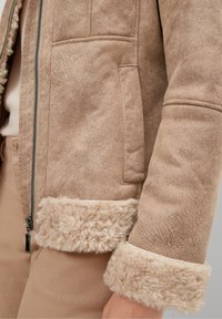 Giacca in faux suede color tan con polsini e colletto beige morbidi e pelosi, dotata di zip frontale e tasche laterali. La texture è liscia e soffice.