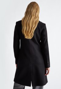 Cappotto nero su misura con maniche lunghe, forma affusolata e uno spacco posteriore. Presenta dettagli con bottoni sui polsini e una texture di tessuto liscia.