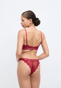 Red lace lingerie set featuring a bralette with adjustable straps and a matching thong with floral patterns and logo detailing.