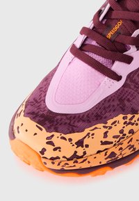 Chaussure de sport violette et rose avec une tige texturée, une semelle intermédiaire tachetée d'orange et une semelle brillante contrastante. Les lacets texturés sont visibles.