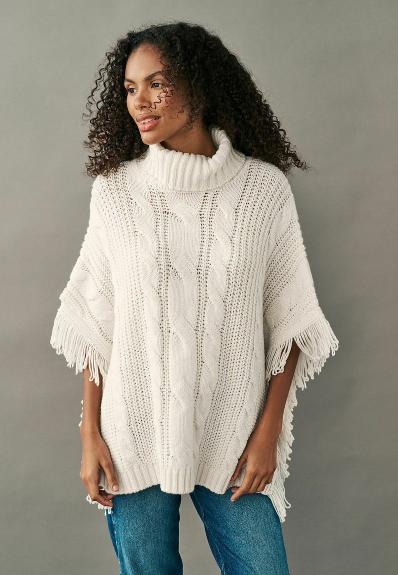 Strickponcho in Weiß mit hohem Kragen. Merkmale sind Zopfmuster, offene Strickabschnitte und fransige Ränder. Kombiniert mit blauen Jeans.