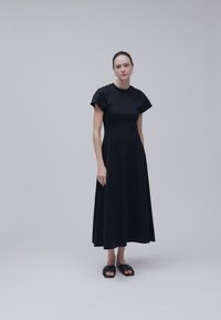 Femme debout sur un fond uni, portant une robe midi noire à manches courtes et des sandales slides noires à bouts ouverts.