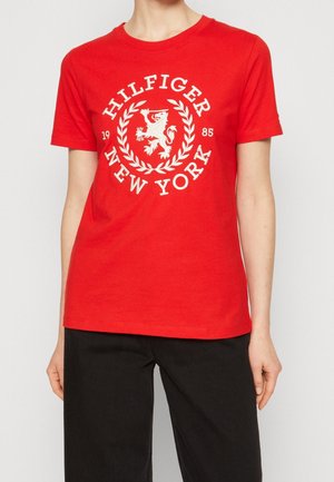 Osoba nosząca czerwony t-shirt z krótkim rękawem z białym tekstem "Hilfiger New York 1985" i emblematem lwa, w połączeniu z czarnymi spodniami.