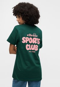 Γυναίκα με κατσαρά μαλλιά που φοράει σκούρο πράσινο μπλουζάκι με ροζ κείμενο "ellesse SPORTS CLUB since 1959" στην πλάτη, σε συνδυασμό με μπλε τζιν.