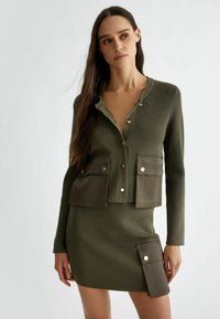 Cardigan a coste in verde oliva con due ampie tasche frontali, abbinato a una gonna coordinata con una tasca laterale e dettagli in hardware argentato.