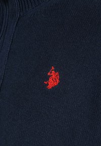 Tissu tricoté bleu marine avec un petit joueur de polo rouge brodé, tenant un maillet levé à cheval.